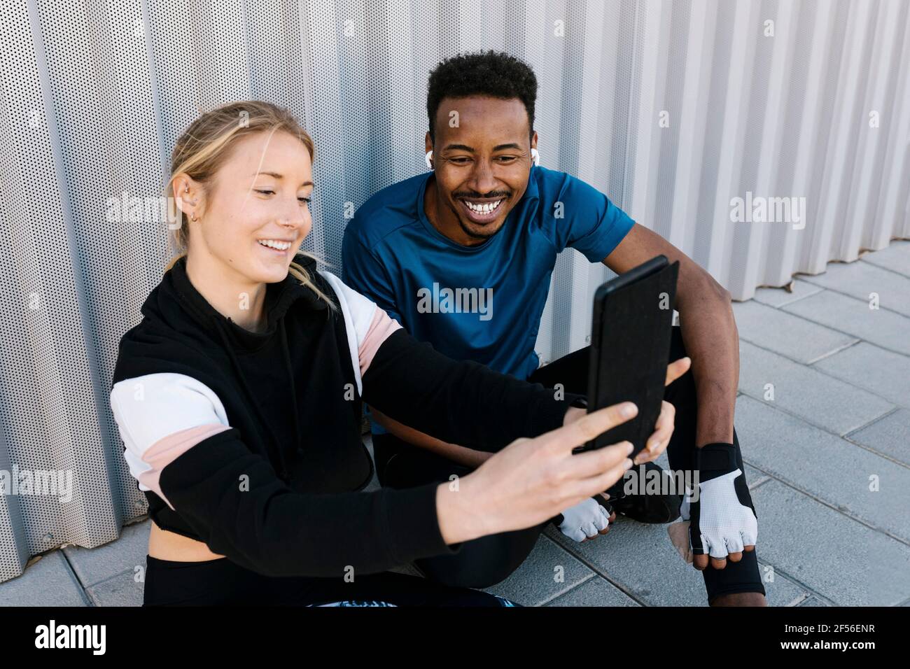 Fröhliche männliche und weibliche Sportlerin, die Selfie über ein digitales Tablet nimmt Auf dem Boden gegen die Wand sitzen Stockfoto