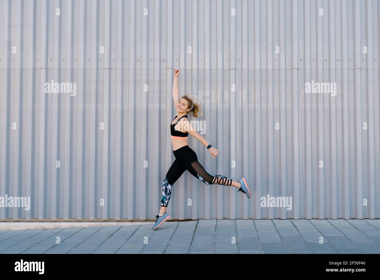 Lächelnde Frau mit erhobenen Armen springt auf Bürgersteig an Wand Stockfoto