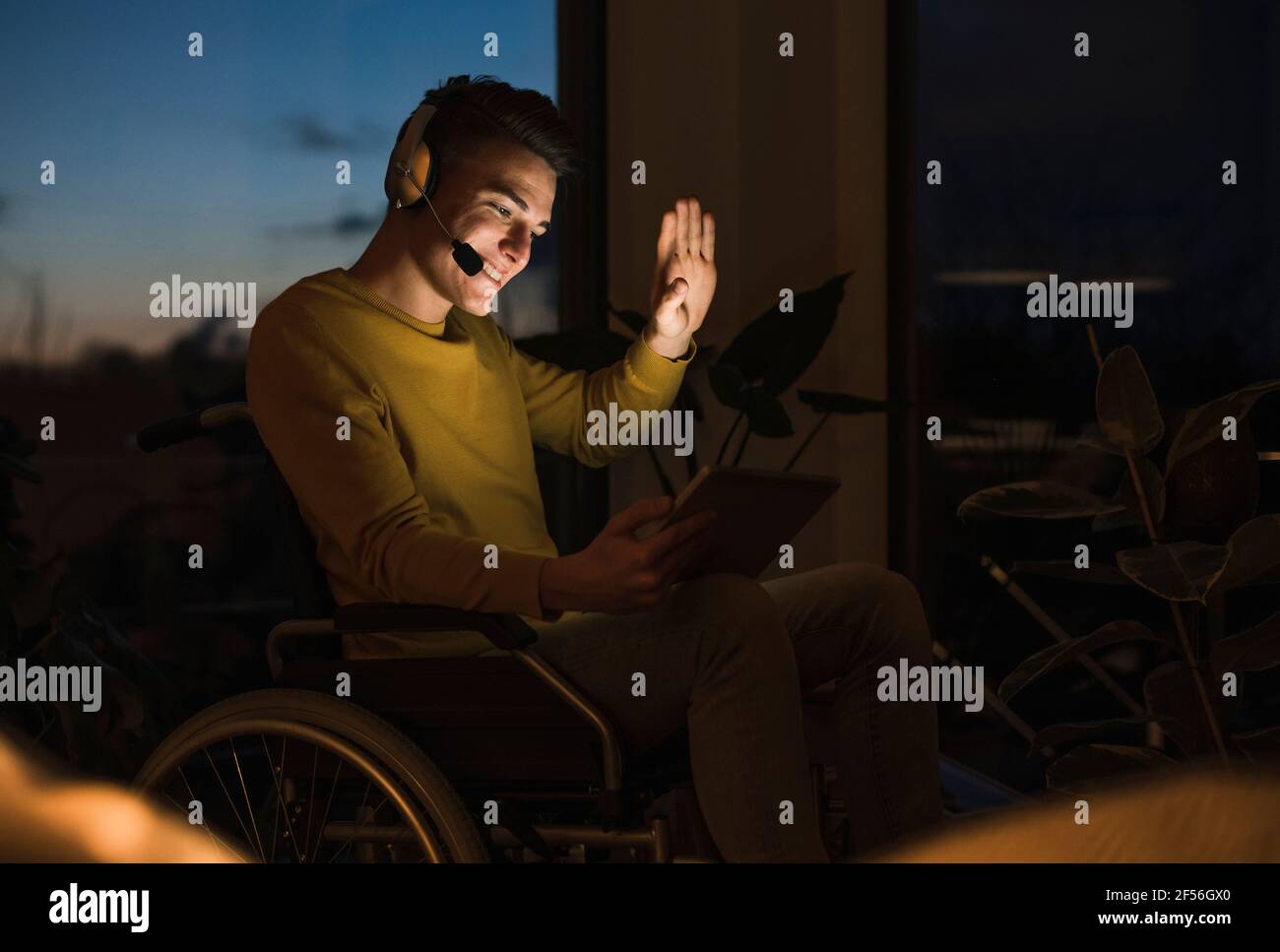 Lächelnder junger Mann im Rollstuhl, der während des Videoanrufs gestikuliert Über ein digitales Tablet, das im Wohnzimmer auf dem Rollstuhl sitzt Stockfoto