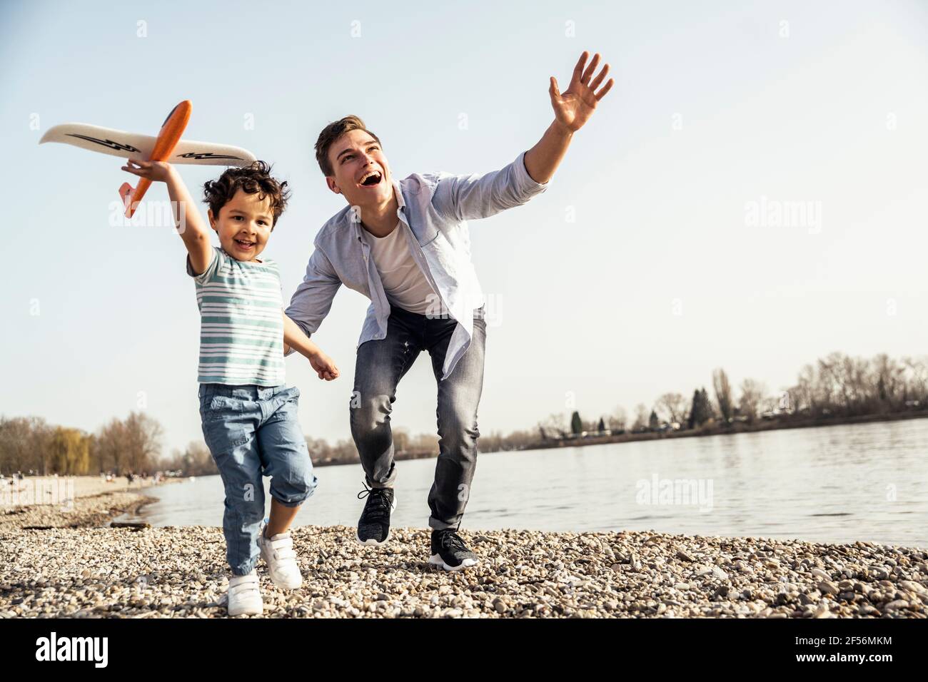 Verspielter Mann und Junge halten Flugzeugspielzeug, während sie vorbei laufen Seeufer an sonnigen Tagen Stockfoto