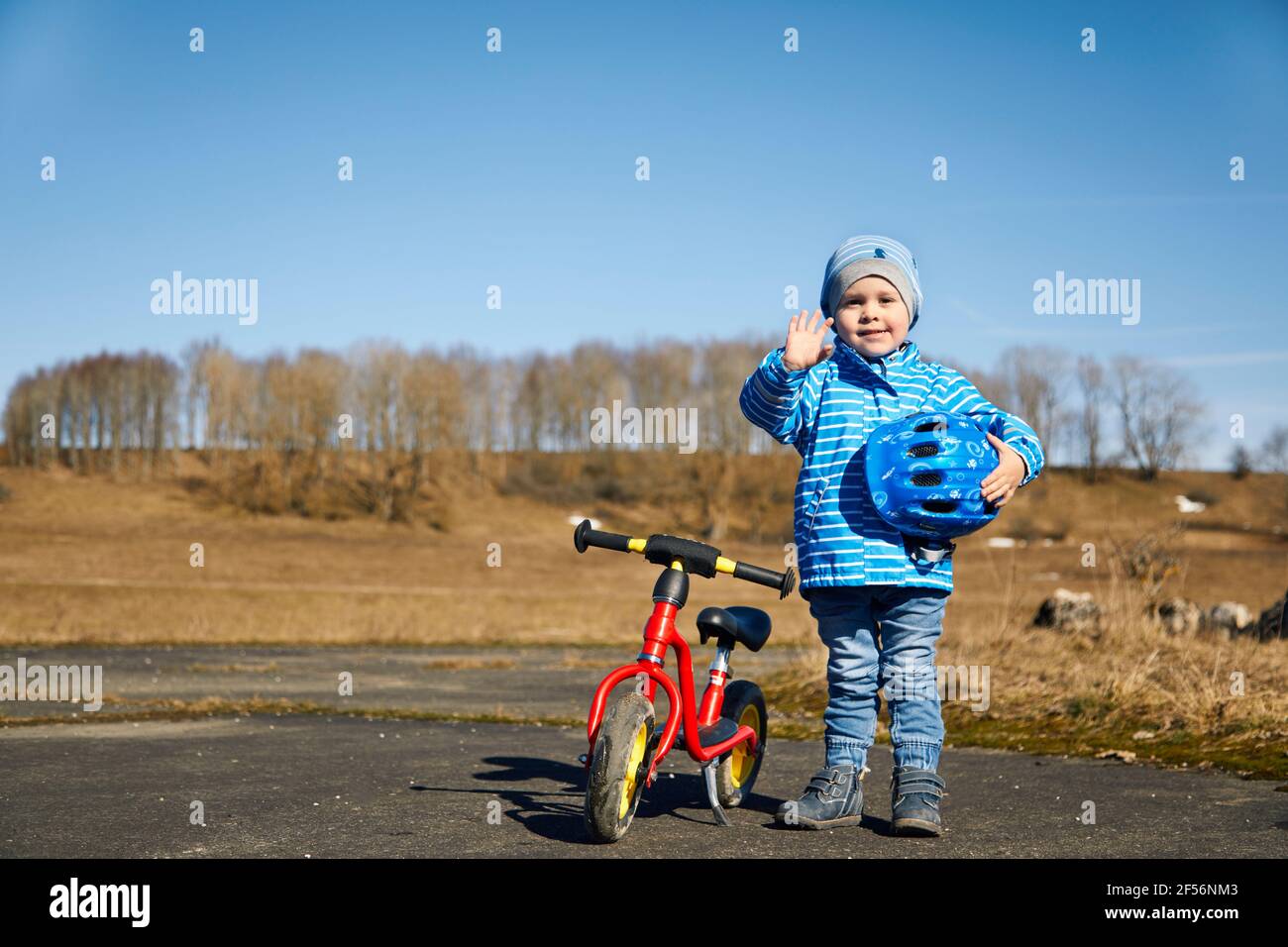 Netter Junge mit Fahrradhelm, der die Hand winkt, während er neben sich steht Laufrad auf der Straße Stockfoto