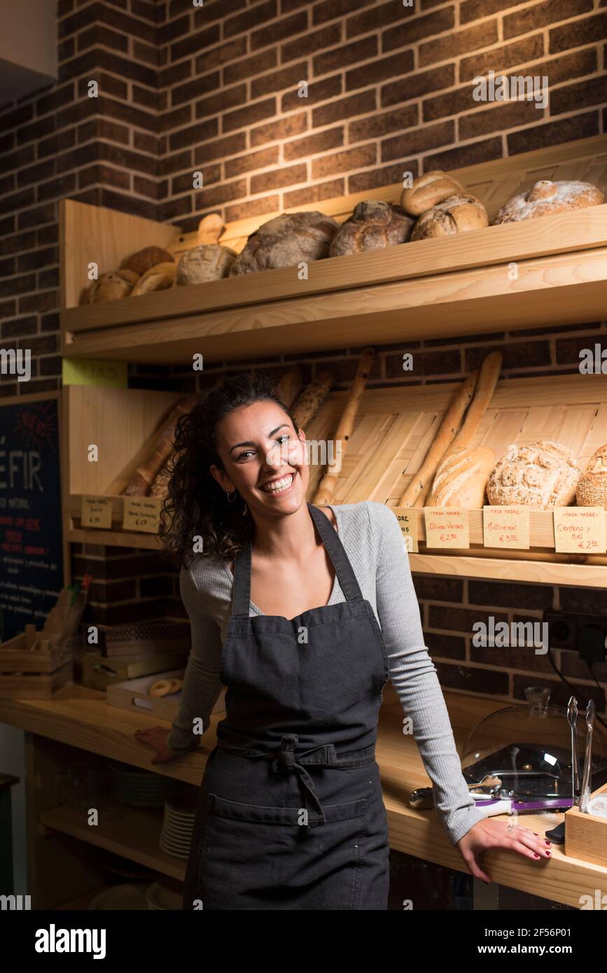 Lächelnder, junger Besitzer, der in einer beleuchteten Bäckerei gegen ein Backwarenregal steht Stockfoto