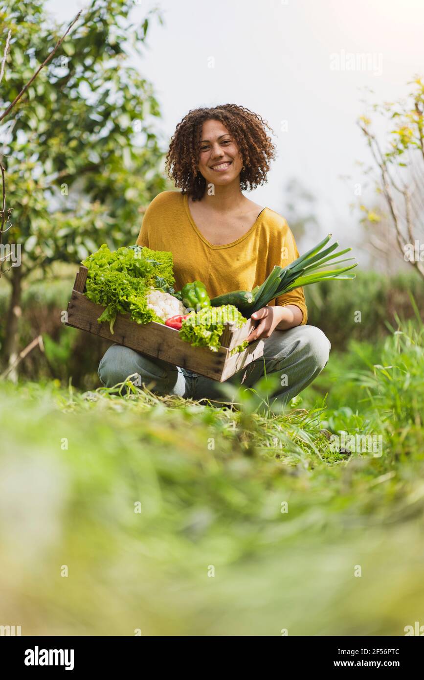 Lächelnde Frau hockt, während sie Blattgemüse gegen den Himmel hält Garten Stockfoto