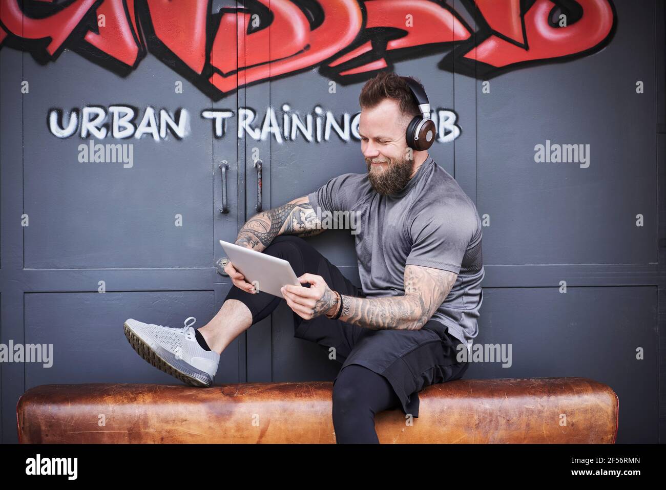 Lächelnder männlicher Athlet, der vor dem Fitnessstudio ein digitales Tablet gegen Graffiti nutzt Tür Stockfoto