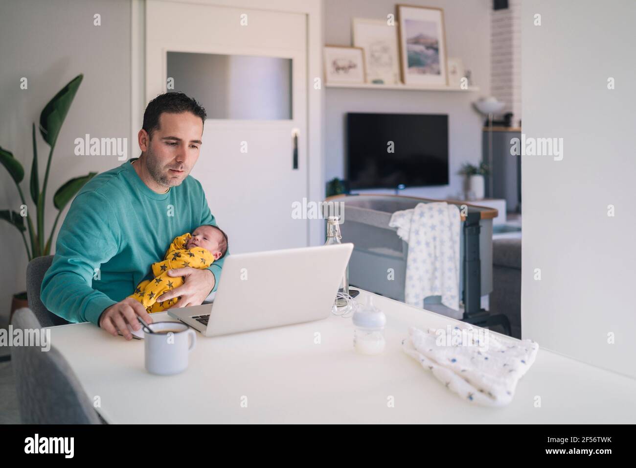 Alleinerziehender Vater, der am Laptop arbeitet, während er seinen Sohn am Schreibtisch trägt Im Heimbüro Stockfoto