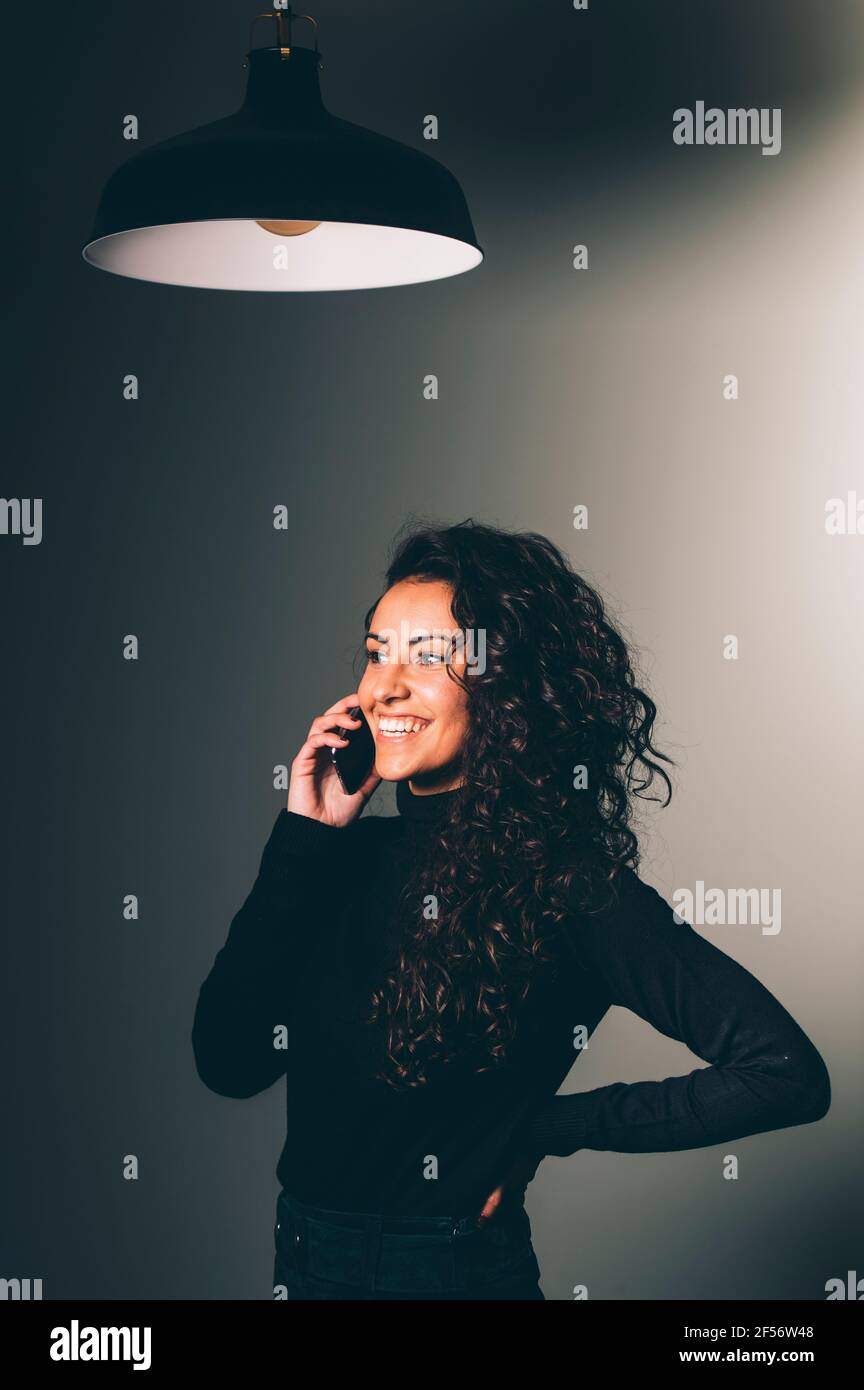 Lächelnde, lockige Geschäftsfrau, die auf dem Mobiltelefon unter dem Pendelleuchte steht Stockfoto