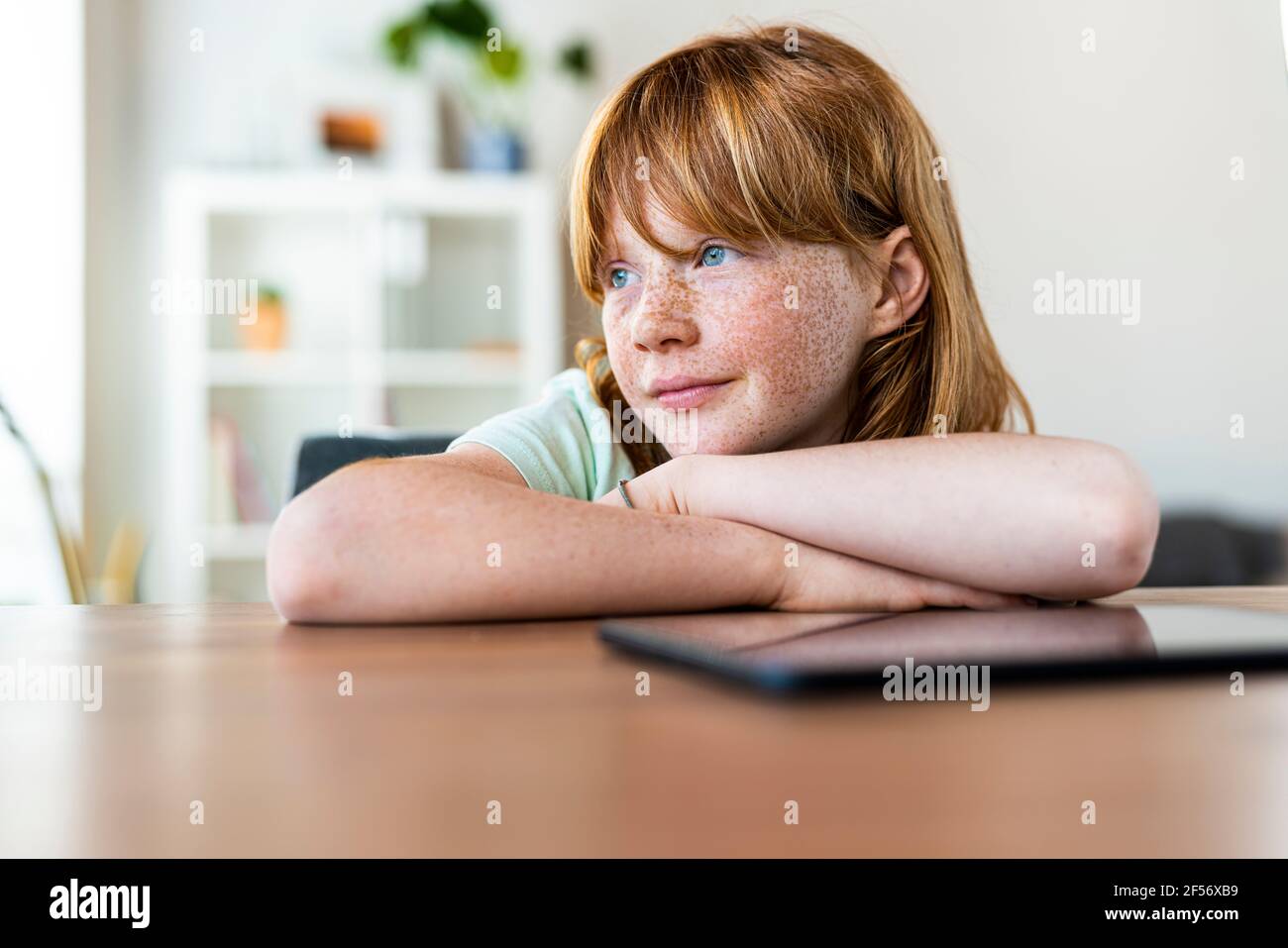 Nachdenkliches rothaariges Mädchen, das wegschaut, während es an einem Tisch sitzt Wohnzimmer Stockfoto