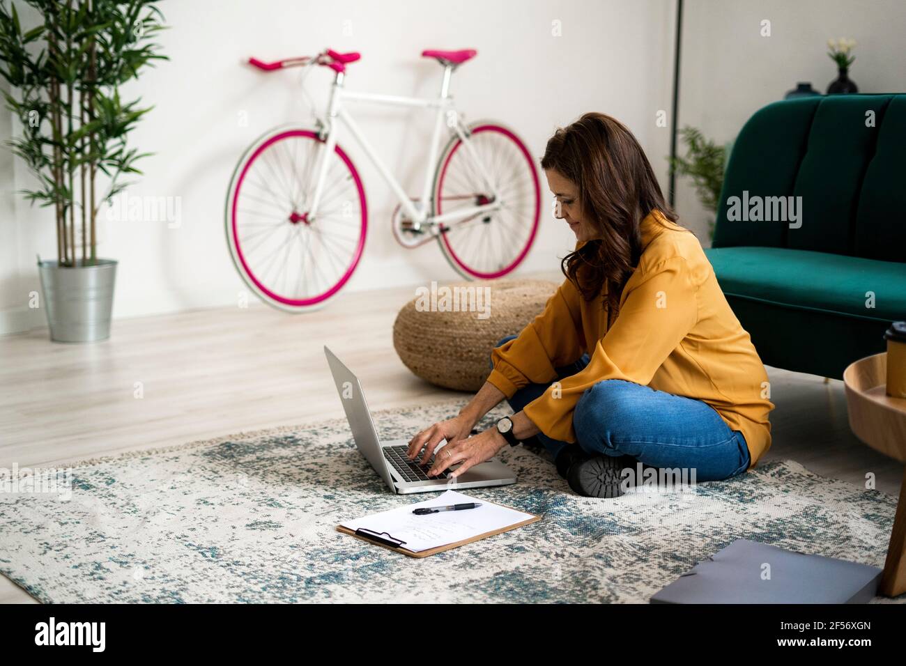 Lächelnde Frau mit Kreuzbeinen, die während des Einsatzes auf dem Boden sitzen Laptop im Wohnzimmer Stockfoto