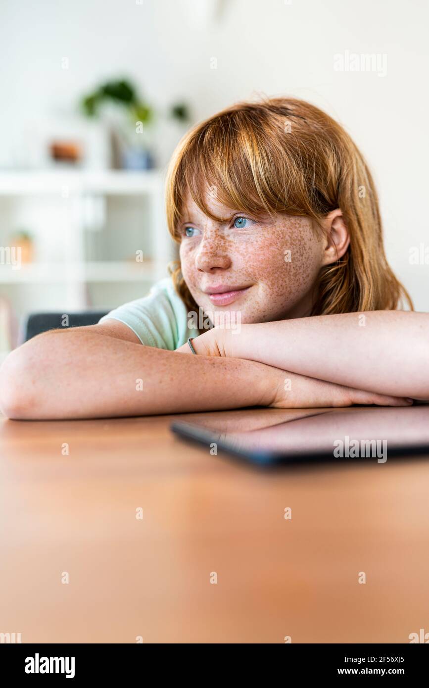 Lächelndes Mädchen mit rothaarig gekreuzten Armen, das beim Sitzen wegschaut Am Tisch im Wohnzimmer Stockfoto
