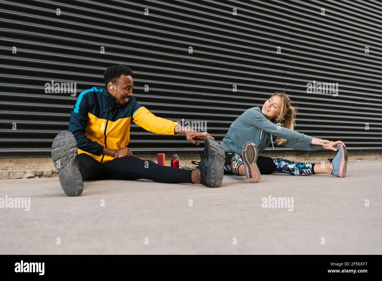 Männliche und weibliche Sportsperson tun Stretching-Übung während des Sitzens auf Boden gegen Rückwand Stockfoto