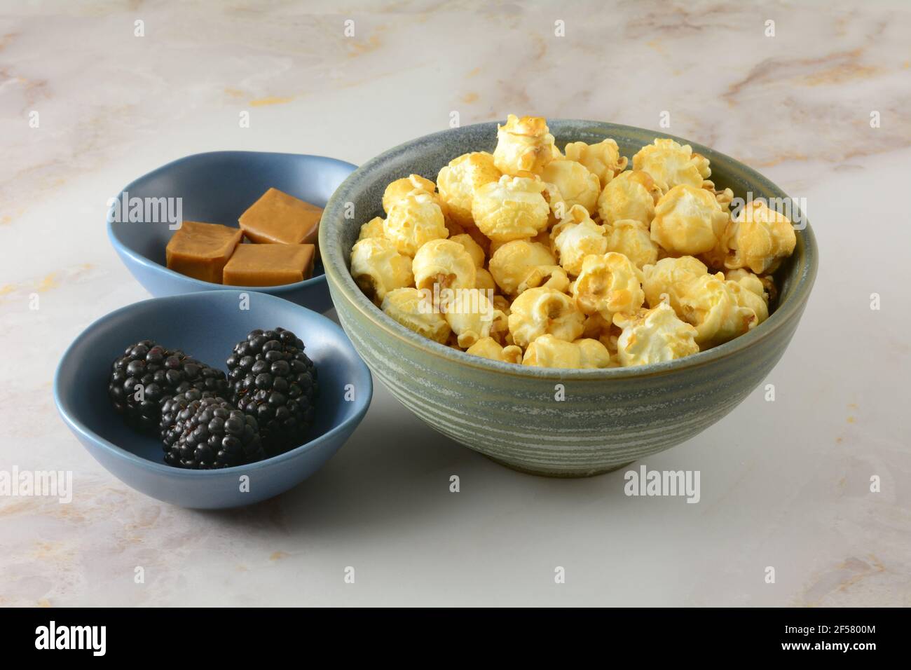 Karamell-Popcorn mit einem Hauch Brombeerlikör im Snack Schüssel und Karamellquadrate und frische Brombeeren in kleiner Zutat Schüsseln Stockfoto