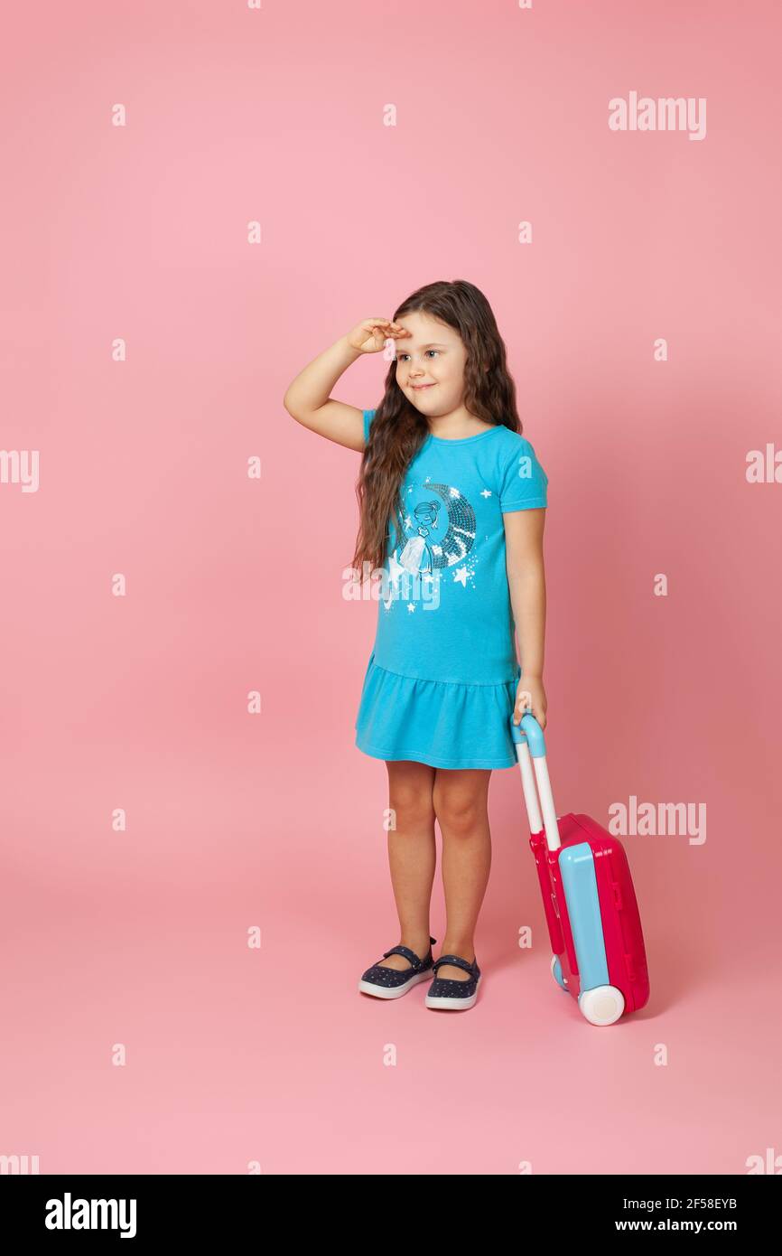 Ganzkörperportrait eines positiven lockigen Mädchens, das einen Koffer trägt und mit der Hand über dem Kopf weit weg schaut, isoliert auf einem rosa Hintergrund Stockfoto