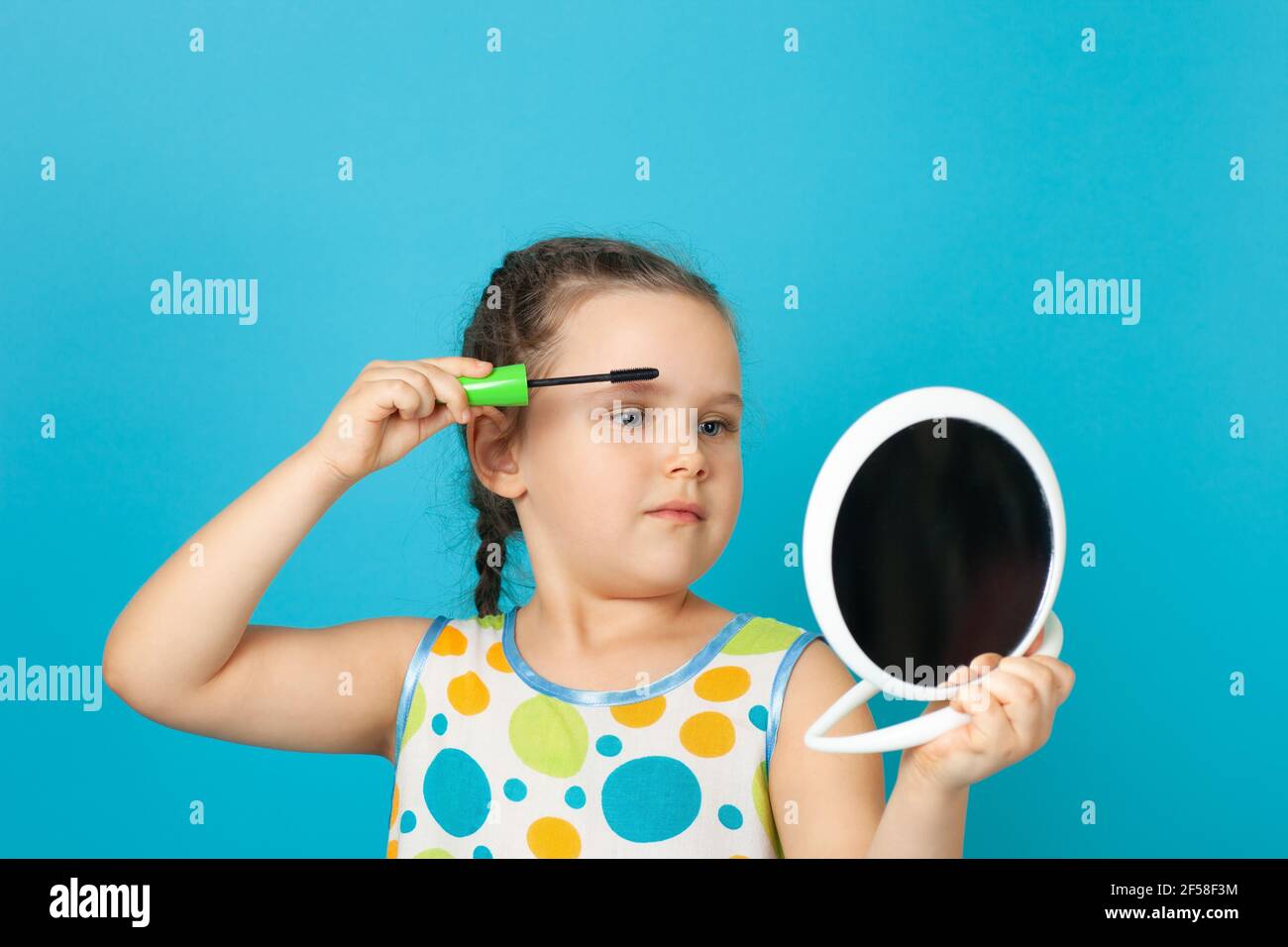 Portrait eines charmanten Mädchens mit Zöpfen, die ihre Wimpern mit Mascara färben und einen weißen Spiegel in der Hand halten, isoliert auf einem blauen Hintergrund Stockfoto