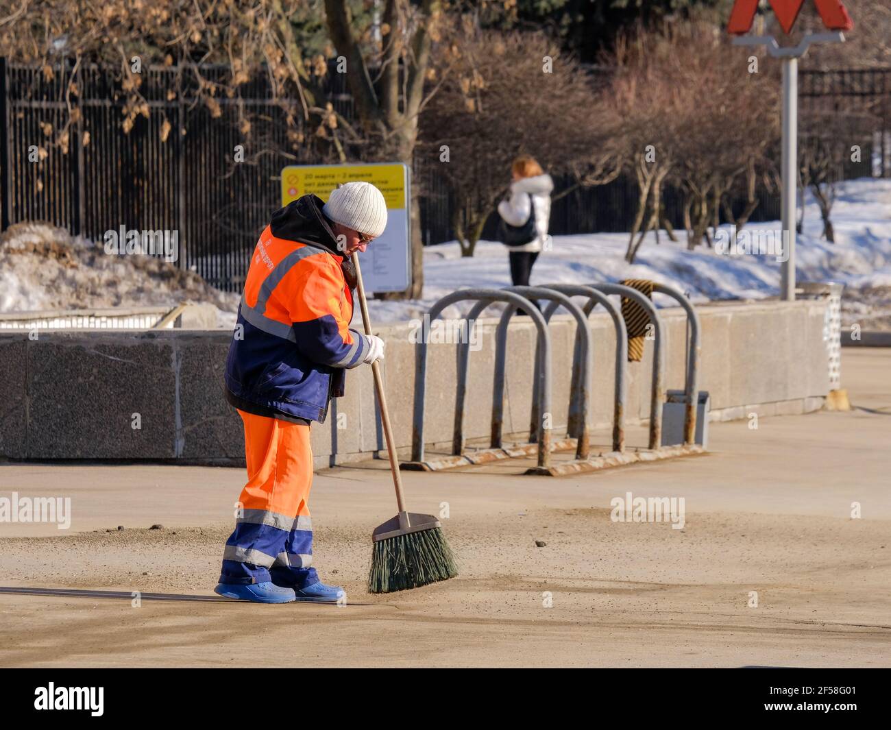 Moskau. Russland. März 24, 2021. Eine Hausmeisterin in orangefarbener Uniform fegt mit einem Besen in der Nähe des Ausgangs der U-Bahn den Bürgersteig. Sonniger Frühlingstag. Der Stockfoto