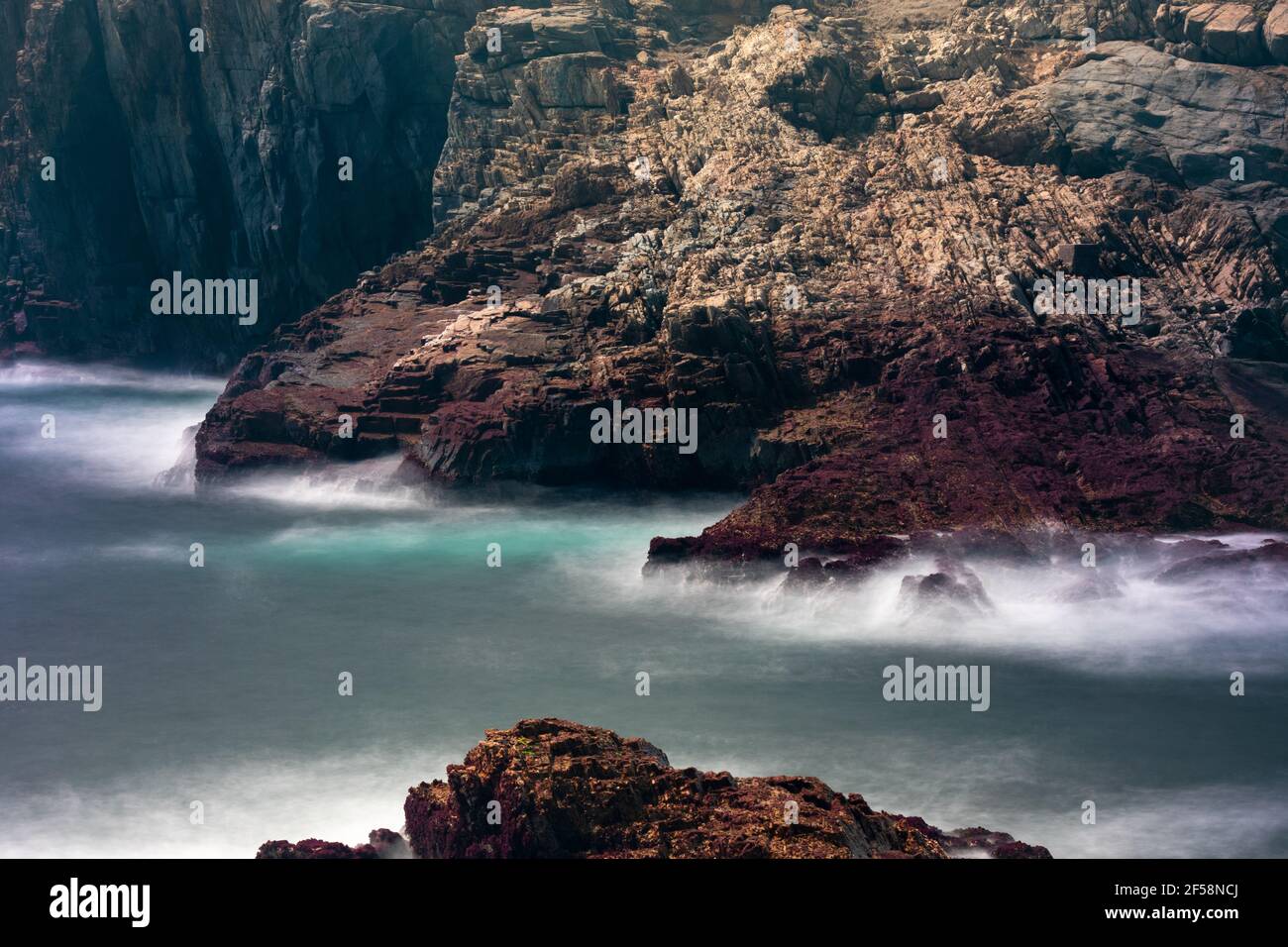 Langzeitaufnahme von Wellen, die auf Felsen brechen und einen seidig glatten Effekt erzielen. Im Hintergrund sind Klippen zu sehen. Stockfoto