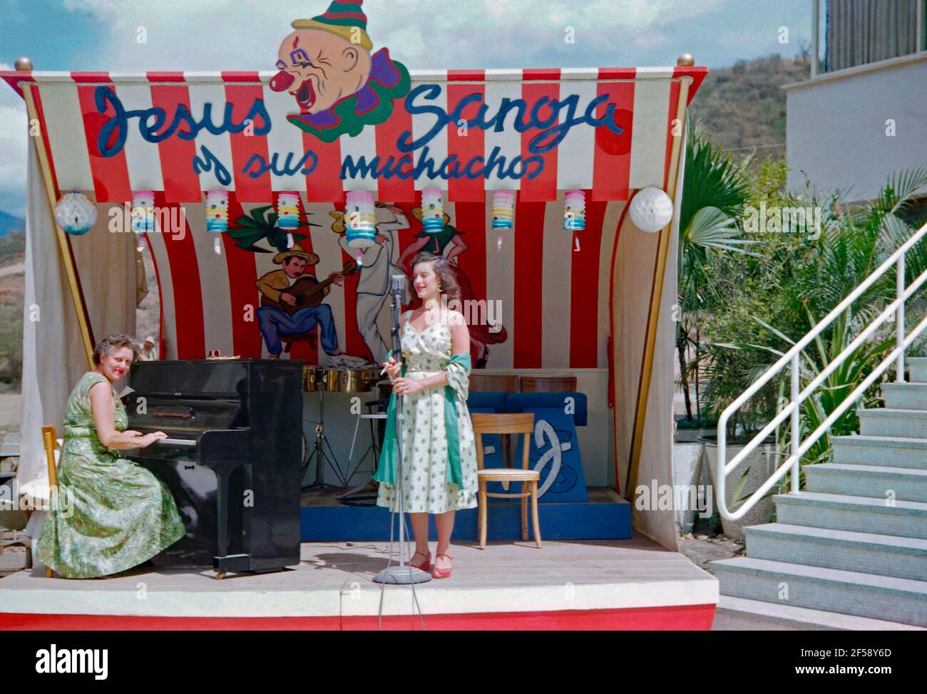 Ein weibliches Musikduo – Sängerin und Pianistin – auf der Bühne im Freien in Venezuela, Südamerika im Jahr 1952. Die Sängerin hält eine Zigarette in der Hand. Die farbenfrohe Kulisse/Baldachin über der Bühne zeigt eine Illustration eines Clowns und hängender Papierlaternen. Es fördert auch die Hauptattraktion, ‘Jesús Sanoja und seine Jungen’. Jesús Chucho Sanoja (1926–1998) war ein venezolanischer Musiker, Pianist, Komponist, Musikdirektor und Arrangeur. Dieses Bild ist von einem alten Amateur 35mm Farbtransparenz - ein Vintage 1950s Foto. Stockfoto