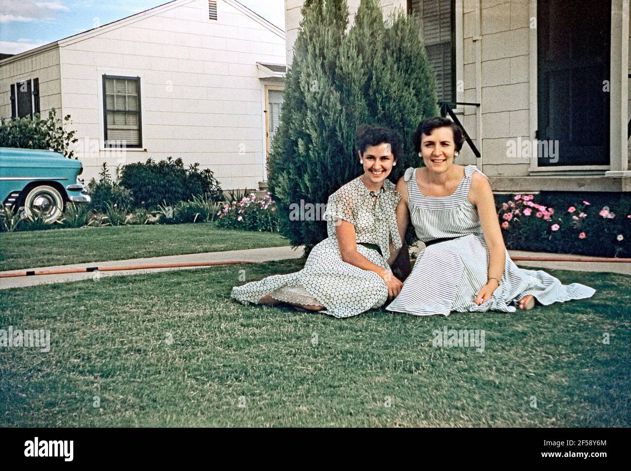 Zwei Frauen posieren auf einem vorderen Rasen neben ihrer S-Zufahrt, USA c. 1960. Die Frauen tragen ihre besten Kleider und lächeln für die Kamera. Dieses Bild stammt von einem alten amerikanischen Amateur Kodak Farbtransparenz - ein Vintage 1950s/1960s Foto. Stockfoto