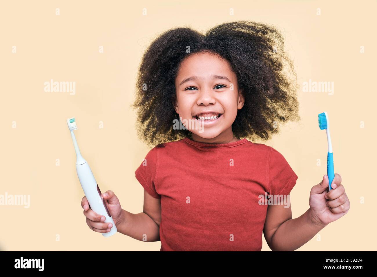 Lächelndes afroamerikanisches kleines Mädchen hält manuelle und elektrische Sonic Zahnbürsten, isoliert auf beigem Hintergrund Stockfoto