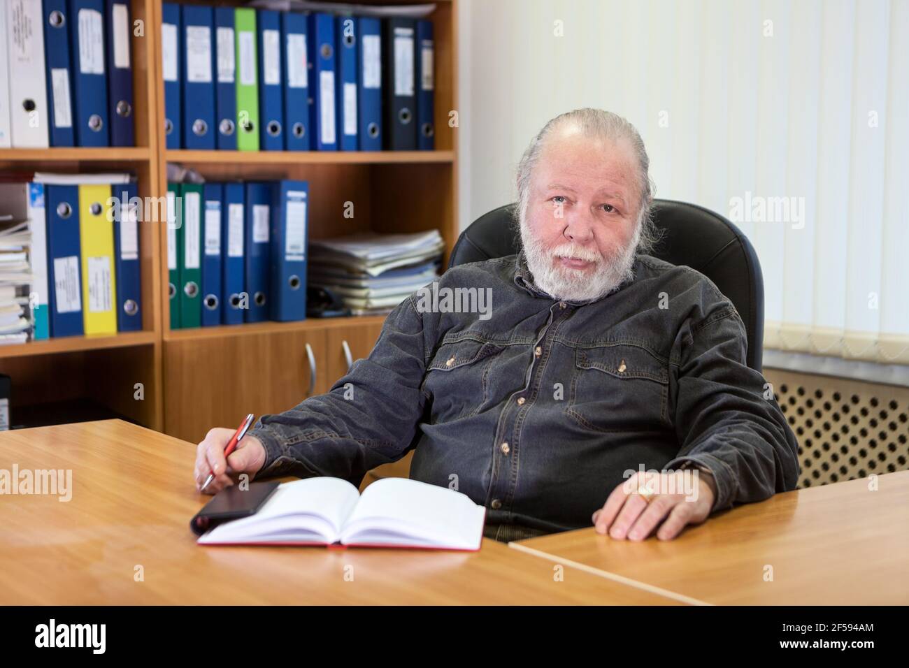 Graues Haar und Bart Professor sitzt am Tisch in seinem Schrank mit einem Notizbuch, kaukasischen älteren Mann Stockfoto