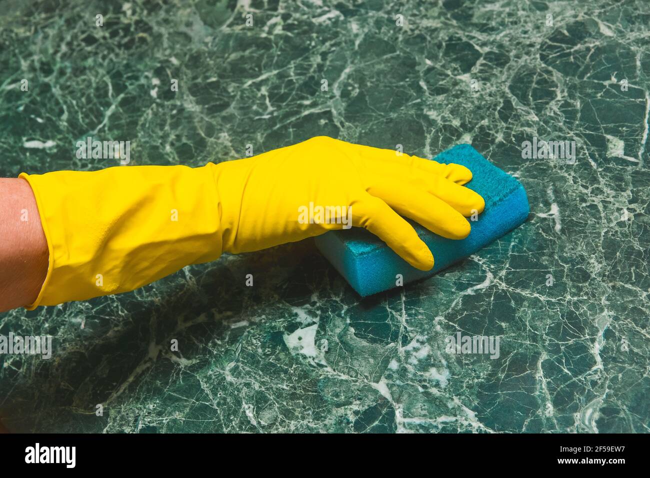 Die Hand der Hausfrau in einem gelben Haushaltshandschuh reibt eine grüne Marmoroberfläche mit einem Schwamm in der Küche. Stockfoto