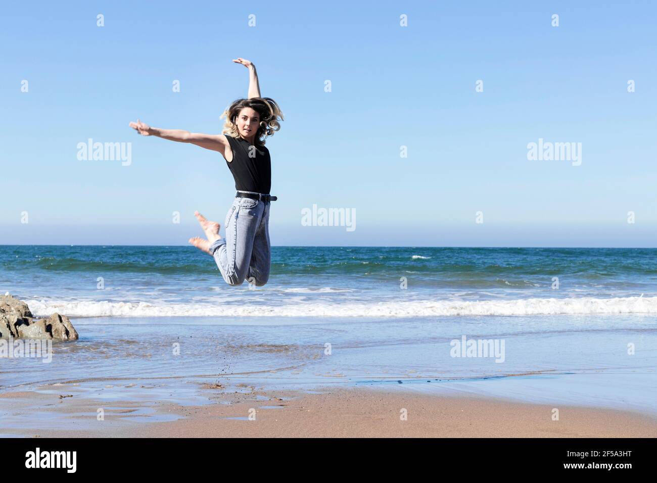 Hübsche junge Frau, die an einem sonnigen blauen Tag am Strand am Meer vor Freude hüpft und Jeans trägt Stockfoto