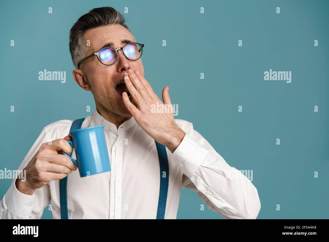 Schläfriger grauhaariger Mann in Brillen gähnend, während Kaffee isoliert trinken Auf blauem Hintergrund Stockfoto