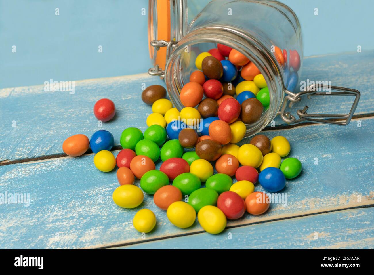 Bunte Süßigkeiten Kugeln im Glas gefallen aus auf blauem Holzhintergrund.Nahaufnahme der bunten Schokolade Süßigkeiten Hintergrund. Stockfoto