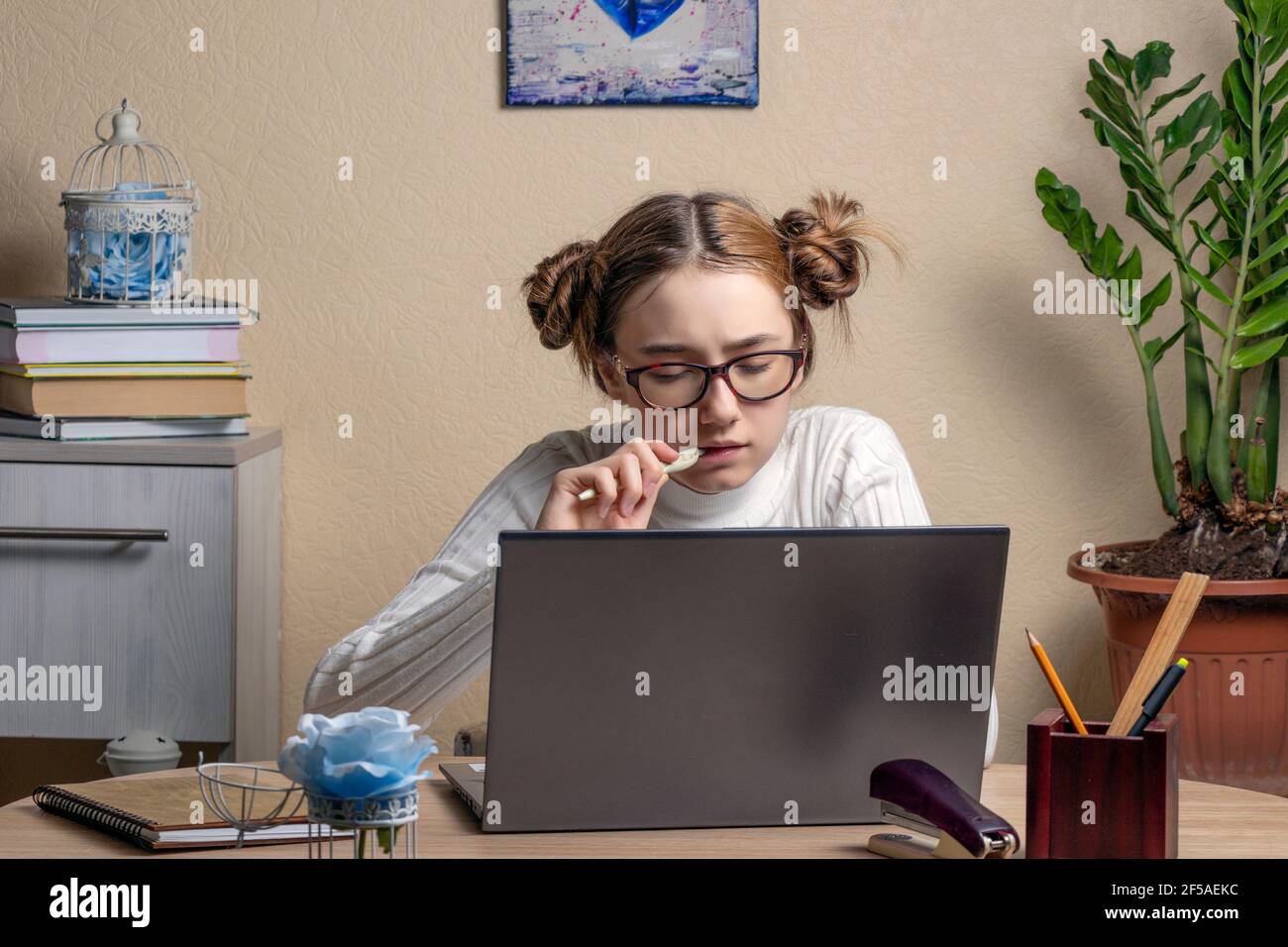 Schöne ernsthafte Teenager-Mädchen in Brille ist online auf einem Laptop verlobt. Ein Teenager-Mädchen starrt aufmerksam auf einen Laptop-Bildschirm und kaut nervös auf einem pe Stockfoto