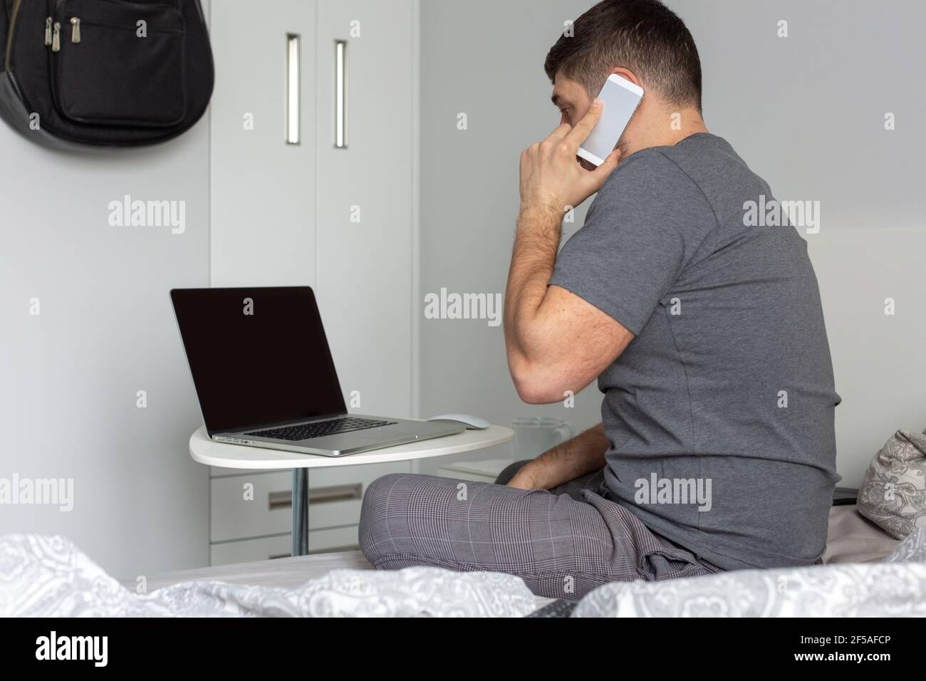 Ein Mann arbeitet hinter einem Laptop aus dem Schlafzimmer, spricht am Telefon während q Stockfoto