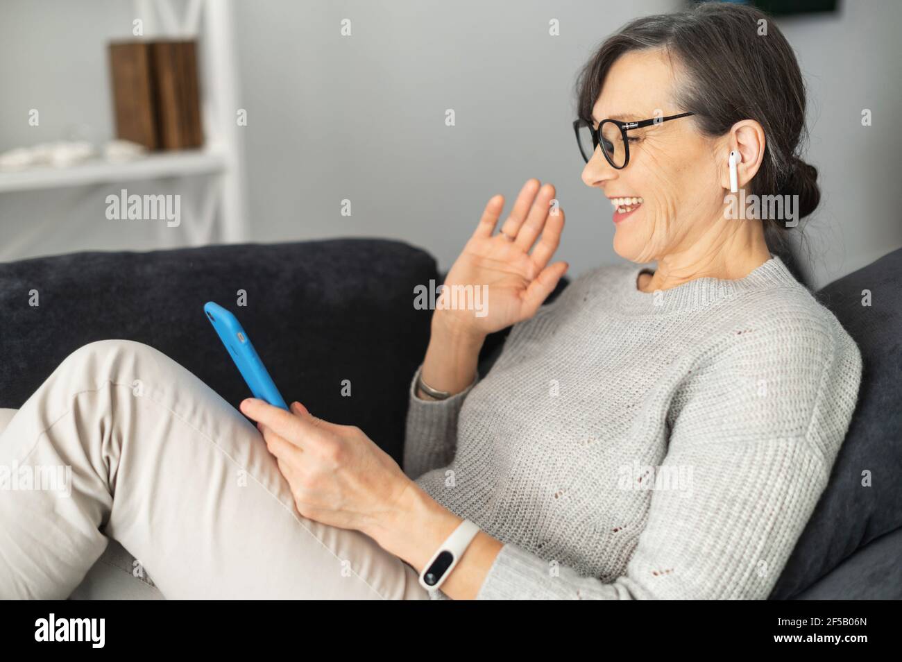 Glückliche ältere Frau, die Videoanruf auf dem Smartphone macht, eine reife  Frau, die auf der Couch liegt, hält das Handy und winkt Hallo in die Webcam,  grüßt Familie oder Freunde auf der