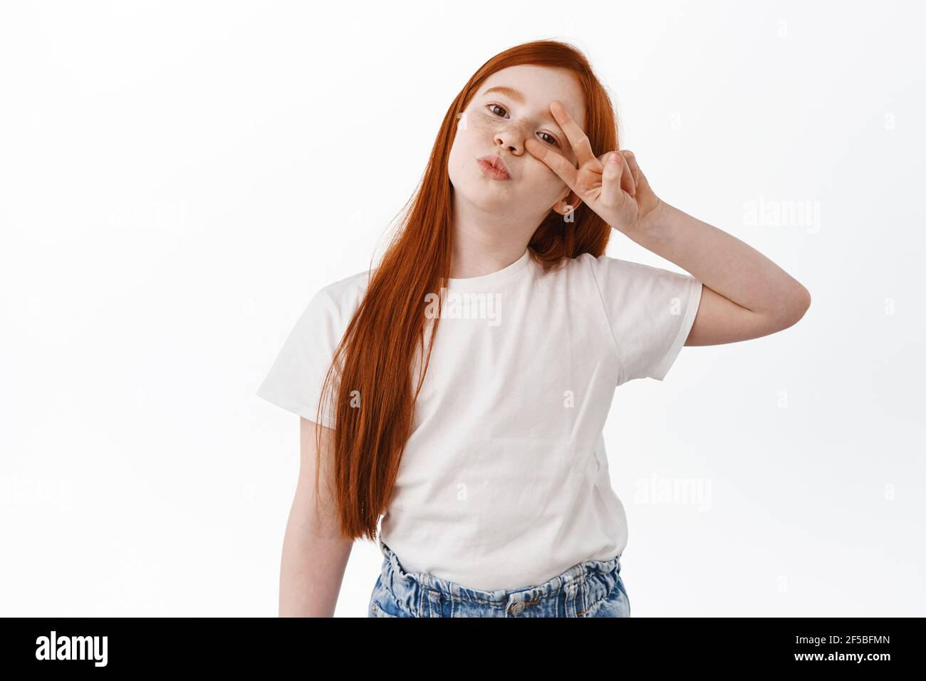 Kinder und Kinder. Schöne kleine Mädchen mit roten Haaren und Sommersprossen machen küssende Lippen und zeigt Friedenszeichen in der Nähe Auge, posiert niedlich gegen weiß Stockfoto