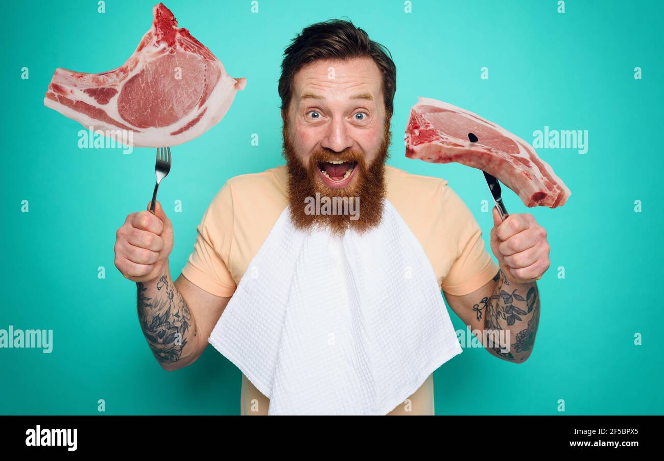 Mann mit Tattoos ist bereit, Fleisch mit Besteck in der Hand zu essen. Cyan Hintergrund Stockfoto