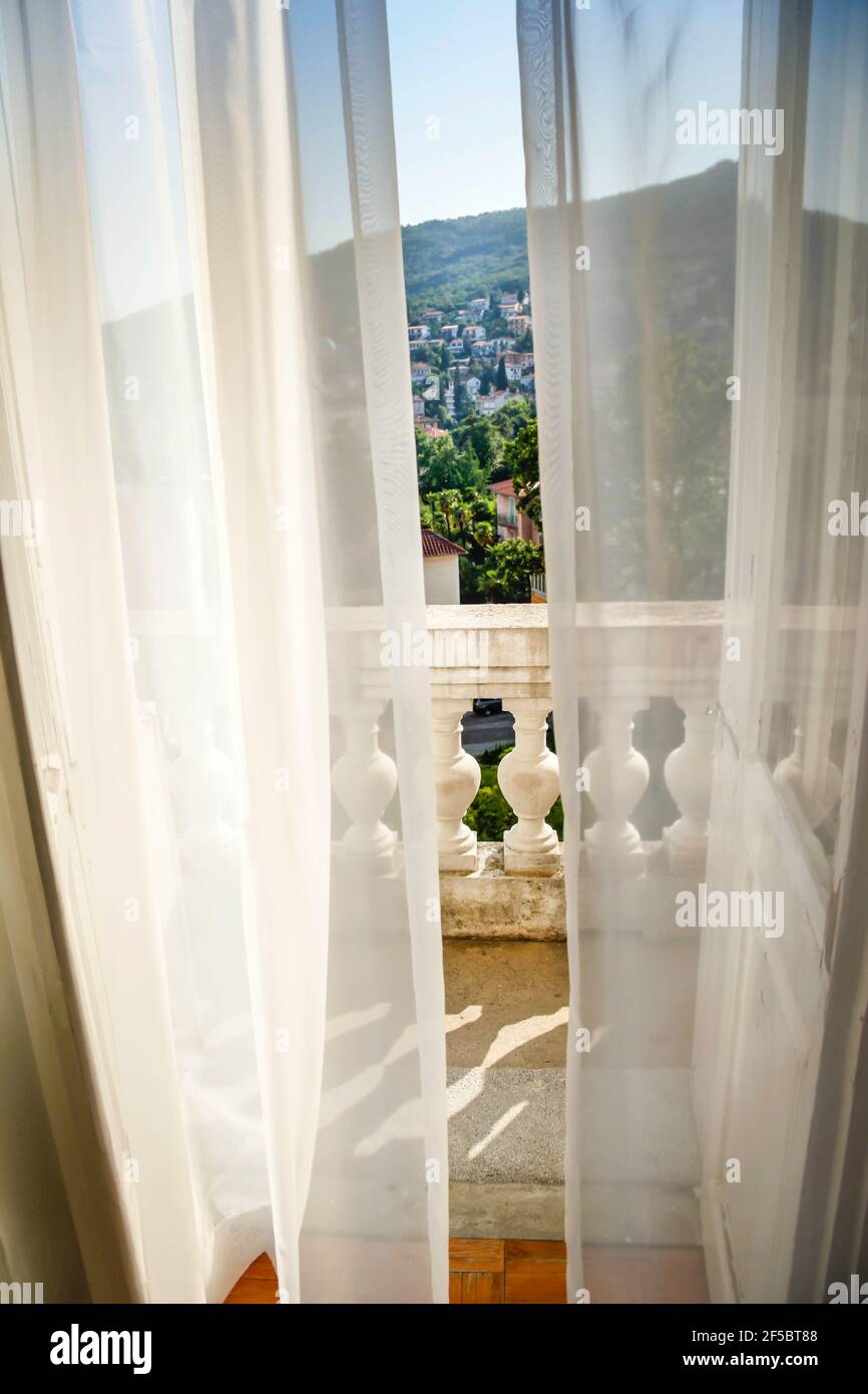 Ein leichter Wind bläst bodenlange Gardinen in einem Romantische Szene von einem offenen Satz von französischen Türen auf Ein Balkon mit einem Blick auf die Vorberge und Häuser Stockfoto