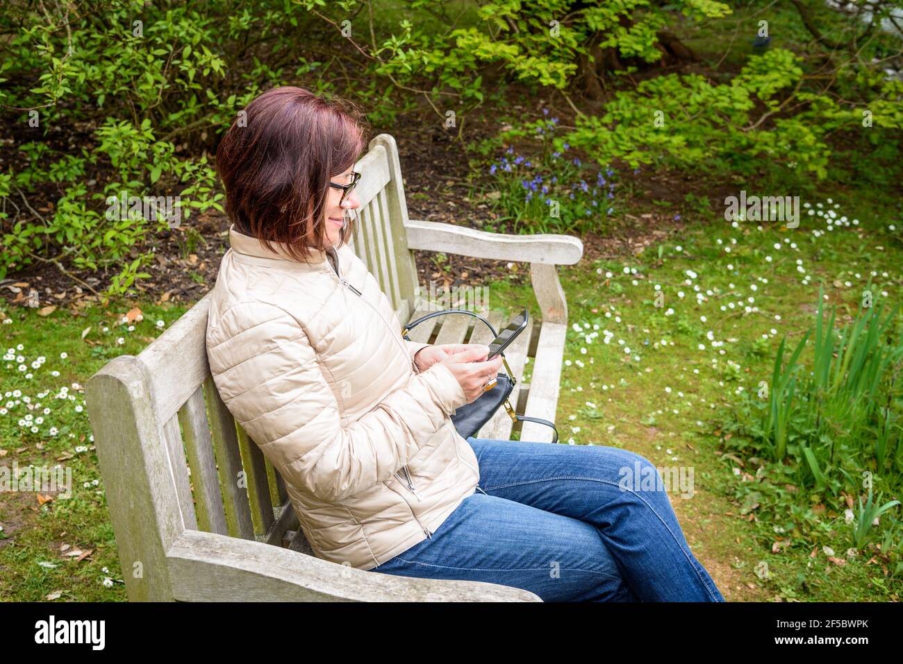 Einsame Frau mit ihrem Smartphone, während sie auf einer Bank sitzt In einem öffentlichen Park im Frühjahr Stockfoto
