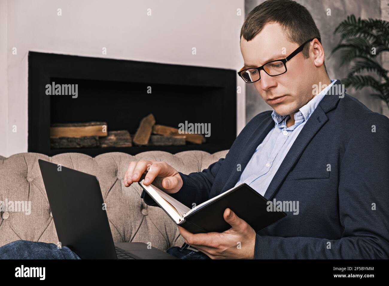 Junge erfolgreiche Mann in Anzug und Brille arbeitet mit Notebook und Laptop sitzen in großen geräumigen Büro auf Sofa vor dem Hintergrund des Kamins. Stockfoto