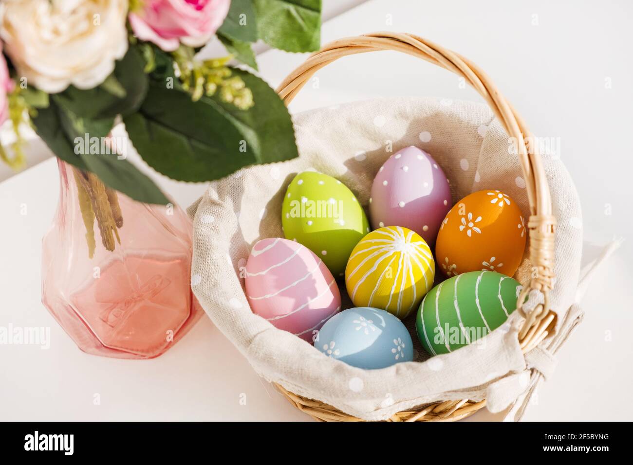 Ostern bunte Eier in busket auf weißem Holztisch. Bouquet von Rosenblüten auf dem Hintergrund. Frohe Ostern. Frühlingsstimmung. Stockfoto