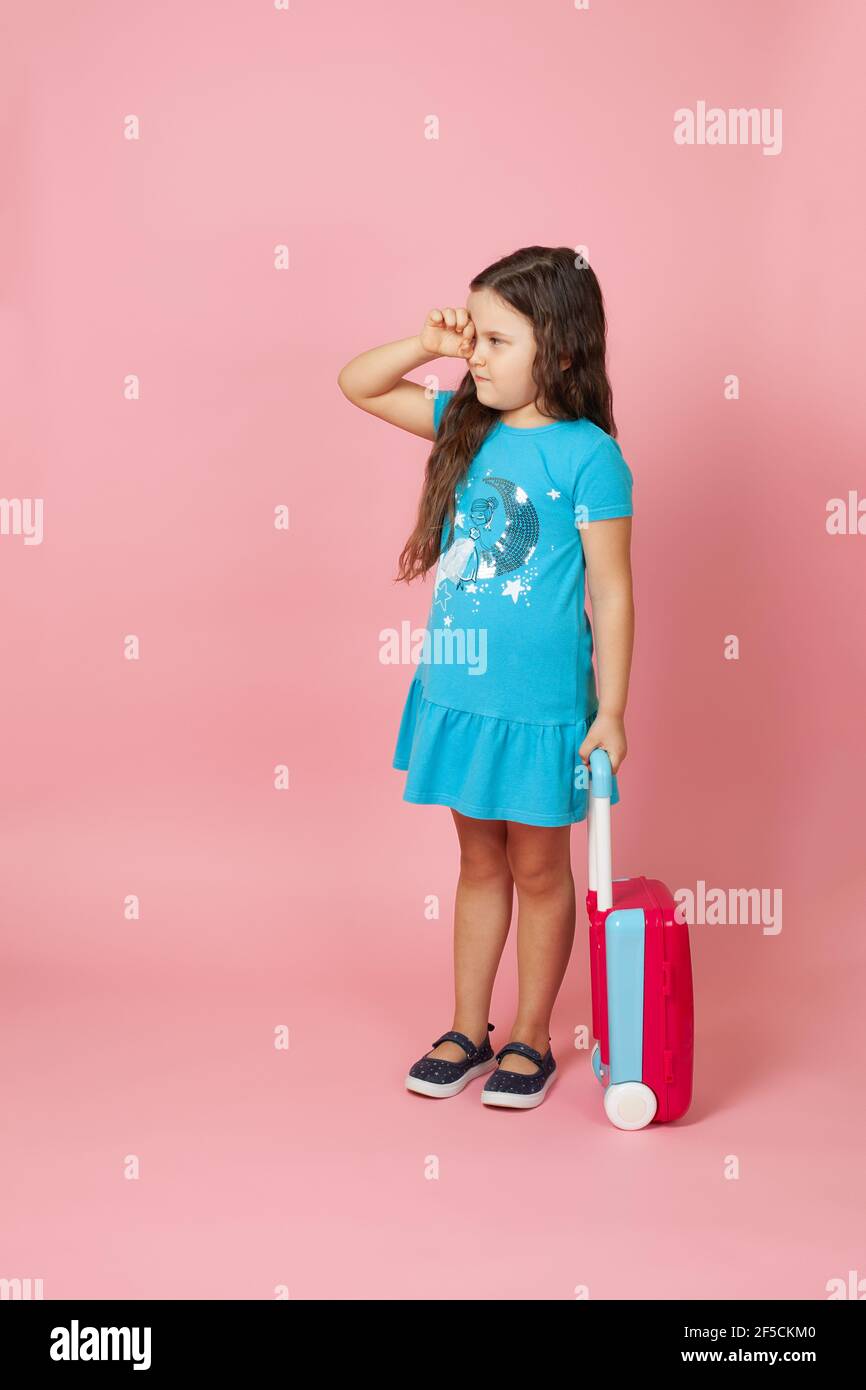 Ganzkörperportrait eines langhaarigen Mädchens, das einen Koffer trägt, mit einem Auge, das durch ihre Finger schaut, isoliert auf einem rosa Hintergrund Stockfoto
