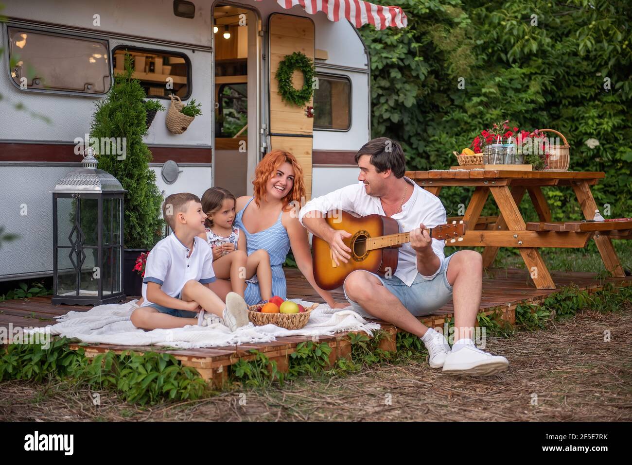 Junger Vater spielt Gitarre, rothaarige Mutter mit Kindern singt Lieder. Glückliche Familie auf Picknick, Camping mit Anhänger LKW in der Natur außerhalb der Stadt Stockfoto