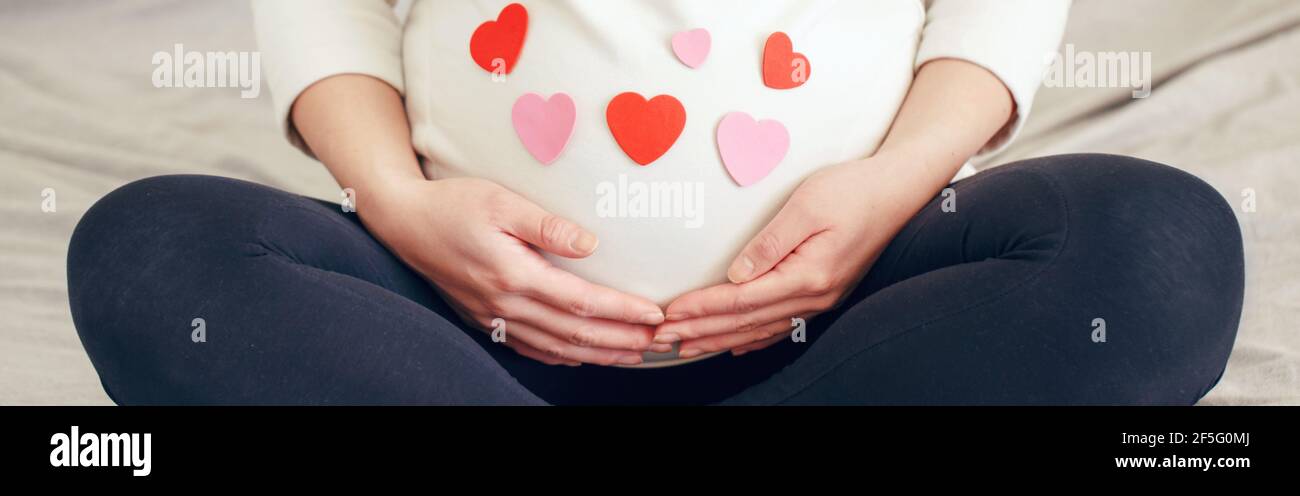 Nahaufnahme einer schwangeren Frau in weißen Kleidern, die auf dem Bett sitzen und den Bauch berühren. Schwangere Bauch mit rot rosa Papier Herzen bedeckt. Junge Dame erwartet Stockfoto