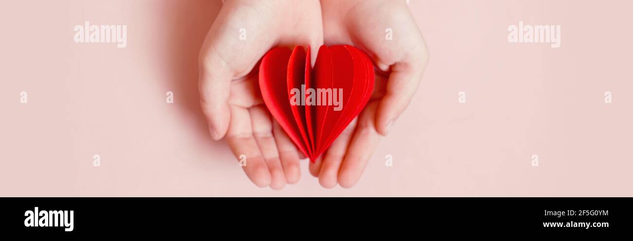 Nahaufnahme von Kind Kind Hände halten roten Papier gefaltet Herz auf hellrosa Hintergrund. Valentinstag Urlaub. Unterstützung, Pflege und Liebeskonzepte. Stockfoto