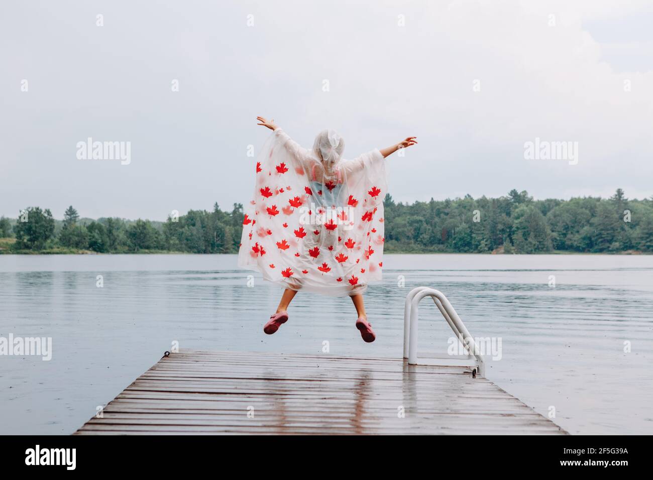Mädchen Kind im Regen Poncho mit roten Ahornblättern Springen auf hölzernen See Dock. Kanada Tag Urlaub. Kind, das im Freien bei Regen die Arme hochhebt. Stockfoto