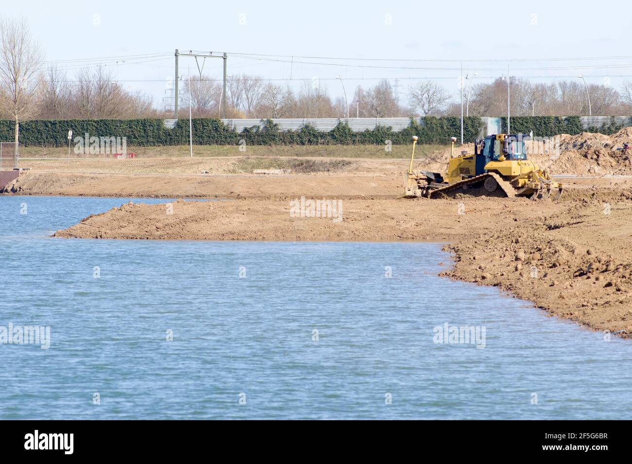 Große gelbe Bulldozer bewegt Boden an einem Kai von Ein See mit einer Eisenbahn im Hintergrund Stockfoto