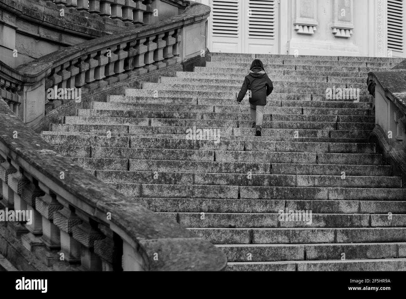 Unbekanntes kleines Mädchen, moderne einsame Prinzessin, die in Europa über majestätische gekrümmte Treppen eines leeren Schlosses geht. Schwarz-Weiß, niedriger Winkel Stockfoto