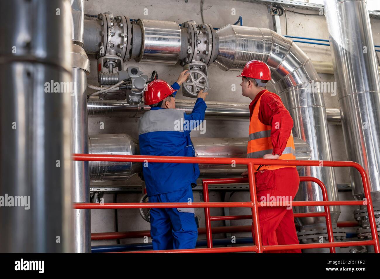 Young Worker in Red Coveralls und HardHat stehen neben Maintenance Worker in blauer Uniform. Werksinneneinrichtung mit großen Rohrleitungen. Stockfoto