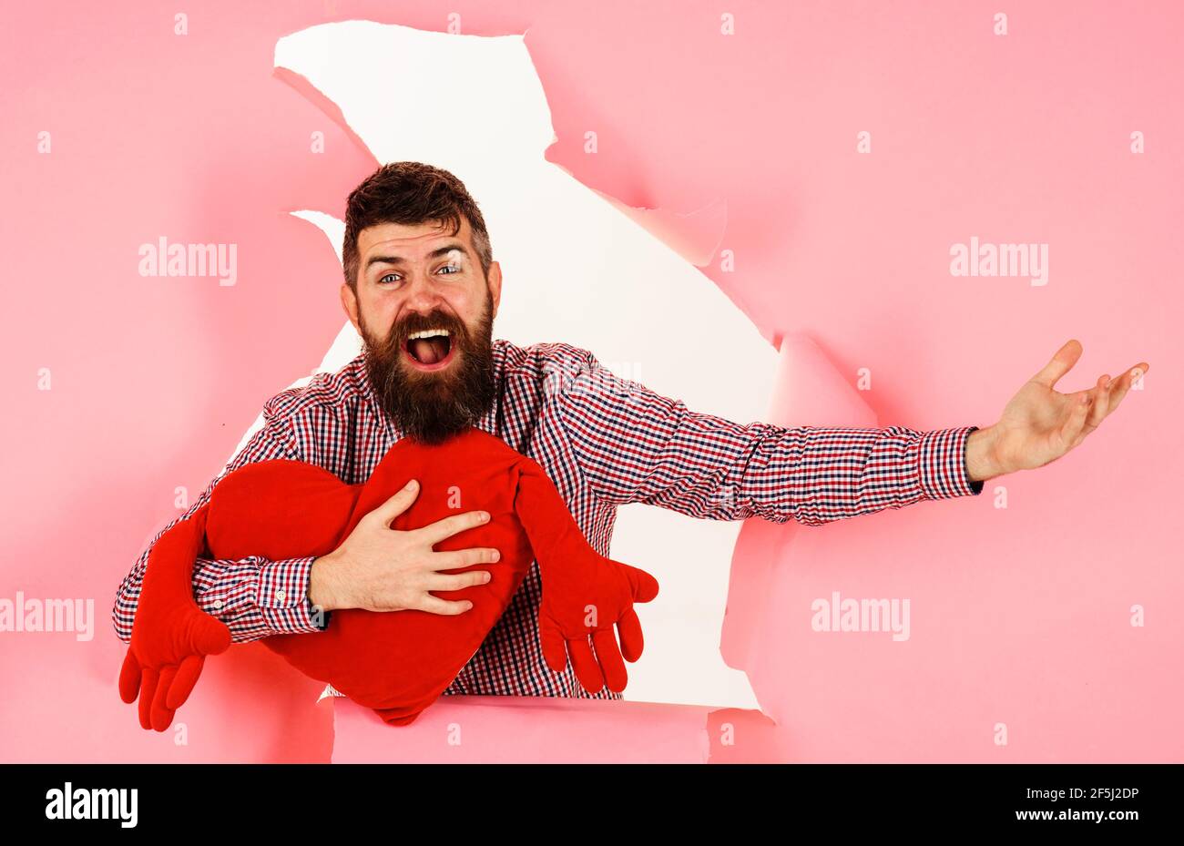 Lächelnder Mann mit herzförmigem Kissen. Bärtiger Kerl mit rotem Herzkissen. Liebe Symbol. Valentinstag-Konzept. Stockfoto