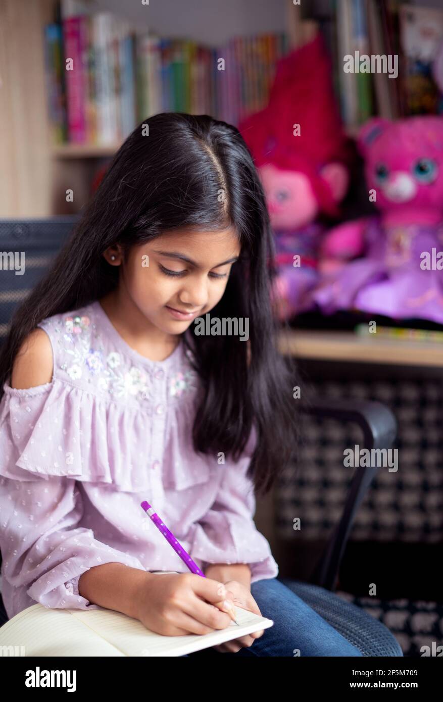 Porträt eines niedlichen indischen Schulmädchen sitzt auf einem Lerntisch und Schreiben eines Tagebuchbuches umgeben von Spielzeug Stockfoto