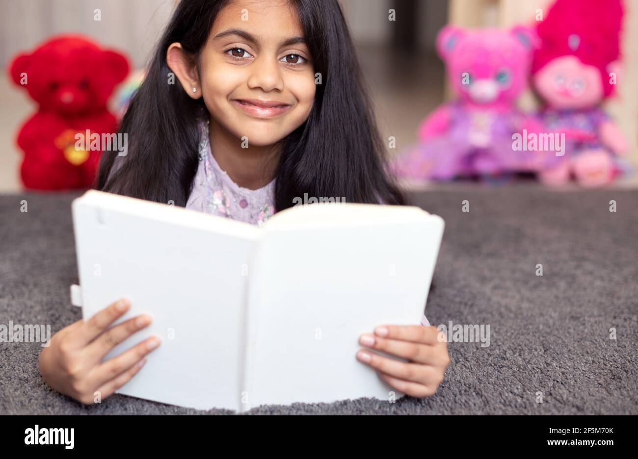 Porträt eines niedlichen indischen Schulmädchen entspannen auf einer Spielmatte, lächeln und ein Tagebuch Buch von Spielzeug umgeben lesen. Stockfoto