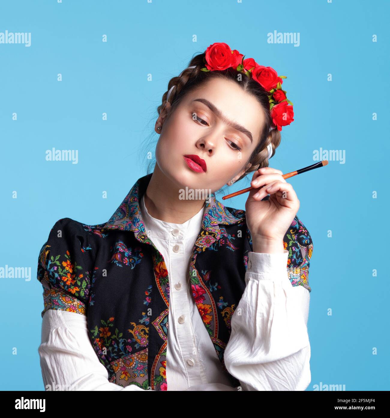 Junge Frau im Bild der mexikanischen Künstlerin Frida mit roten Rosen im Haar. Mit einem Pinsel in den Händen, berühren Augenbrauen. Stockfoto