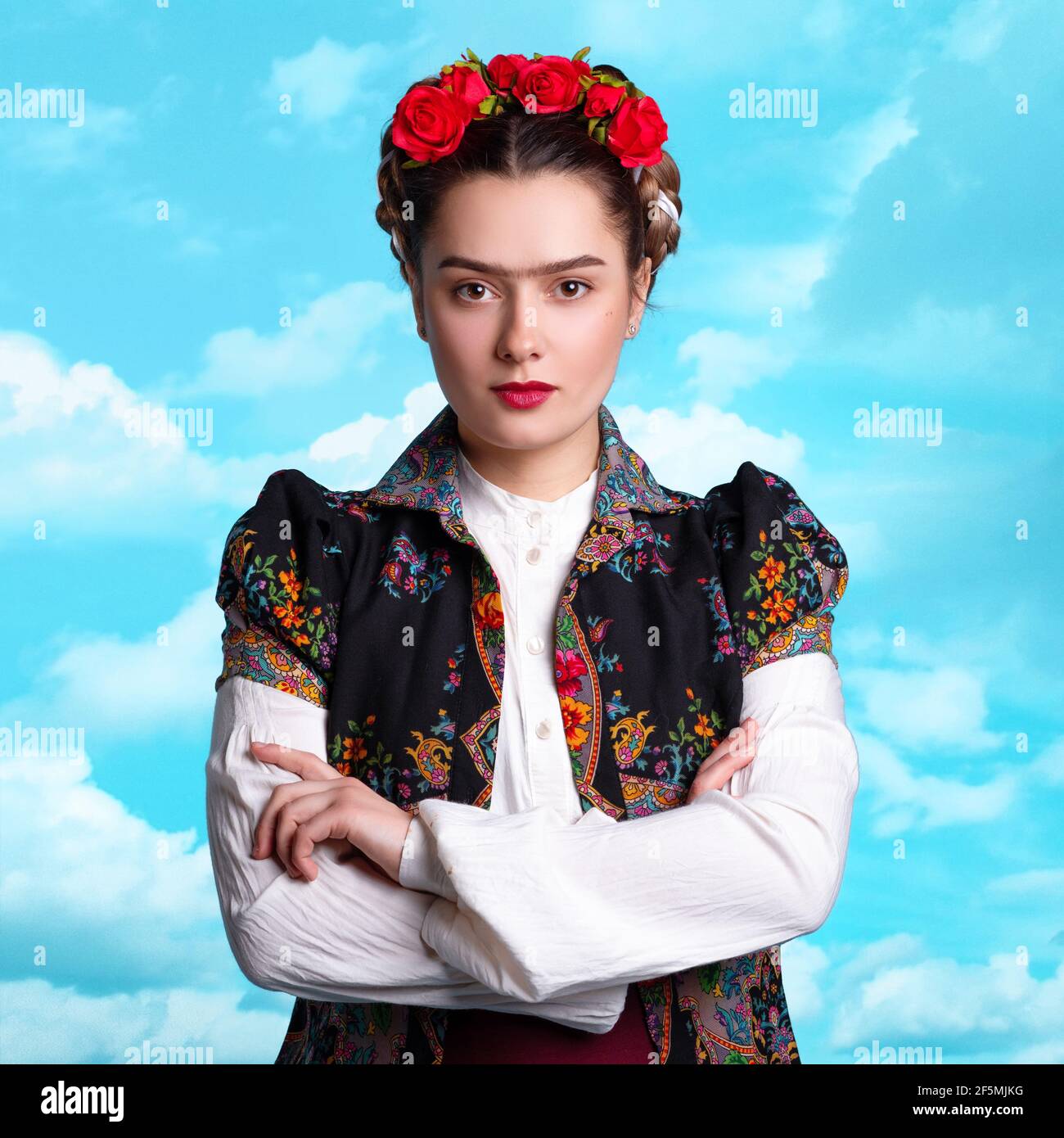 Junge Frau im Bild der mexikanischen Künstlerin Frida mit roten Rosen im Haar. Surrealer Hintergrund mit Himmel. Stockfoto