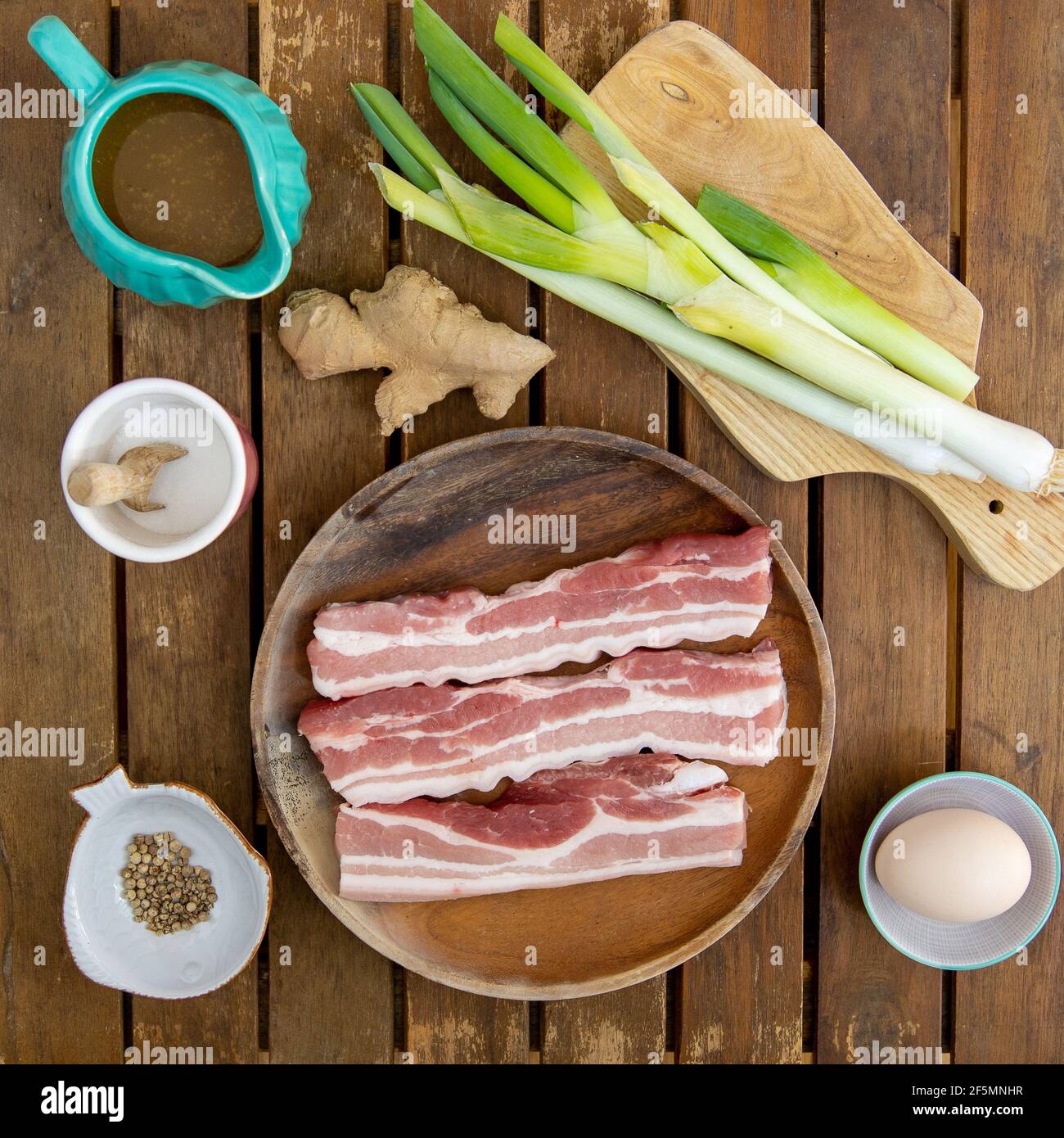 Holztisch mit Schweinefleisch, Frühlingszwiebeln, Ingwer, Brühe, Salz, Weißer Pfeffer, ein Ei und 3 Stück Schweinebauch (Zutaten für chinesische Knödel) Stockfoto